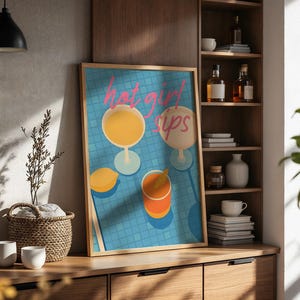 Könnte beinhalten: Ein rechteckiges gerahmtes Wandbild mit einem blauen Fliesenpool-Hintergrund, drei Getränken und einer Zitrone. Der Text hot girl sips ist in rosa Schreibschrift oben aufgedruckt. Das Kunstwerk befindet sich in einem hellen Holzrahmen.