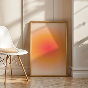 May include: A framed abstract art print with a gradient of orange, pink, and white hues. The artwork is displayed on a wooden floor, leaning against a wall. A white chair with wooden legs and a tan cushion is positioned to the left.