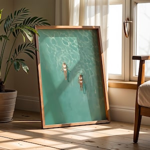 Könnte beinhalten: Ein gerahmter Kunstdruck zeigt zwei Personen, die in türkisfarbenem Wasser schwimmen. Das Kunstwerk befindet sich in einem Holzrahmen und lehnt an einer Wand. Eine Topfpflanze und ein Stuhl befinden sich im Hintergrund. Das Kunstwerk wird auf einem Holzboden ausgestellt.