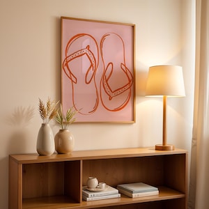 Könnte beinhalten: Ein gerahmter Druck von orangefarbenen Flip-Flops auf rosa Hintergrund hängt über einem Holzregal. Eine Holzlampe mit cremefarbenem Schirm und zwei Vasen mit getrockneten Pflanzen stehen auf dem Regal. Der Raum hat eine neutrale Farbpalette.
