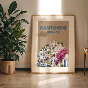 以下が含まれることがあります： サントリーニ島、ギリシャの絵画が描かれた額入りアートプリント。白い建物、青いアクセント、鮮やかなピンクのブーゲンビリアの花が特徴です。ベージュトーンのイラストの上部には、青い文字でSANTORINI GREECEと印刷されています。