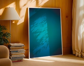 May include: A large framed art print with a teal and blue abstract design, leaning against a wooden wall. The artwork is illuminated by sunlight, casting shadows. A stack of books and a lamp are visible.
