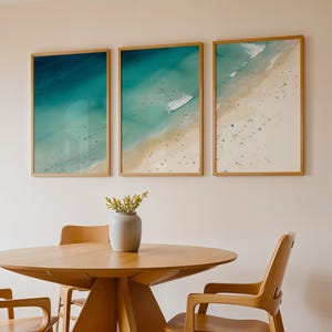 Könnte beinhalten: Drei gerahmte Kunstdrucke mit Strandmotiven in Blau- und Weißtönen hängen über einem Esstisch aus Holz. Der Tisch ist rund mit hellem Holzfinish und wird von passenden Holzstühlen umgeben. Eine Vase mit Grünzeug steht auf dem Tisch.