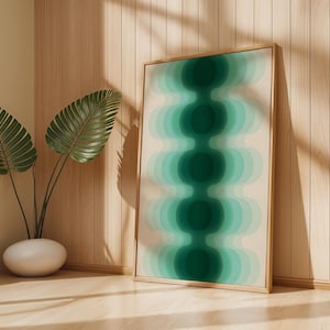 Könnte beinhalten: Ein gerahmter Kunstdruck mit einem abstrakten Design aus geschichteten, abgerundeten Formen in Türkis und Grün auf cremefarbenem Hintergrund. Das Kunstwerk befindet sich in einem hellen Holzrahmen und lehnt an einer Holzwand. Eine Topfpflanze mit großen grünen Blättern steht in der Ecke.