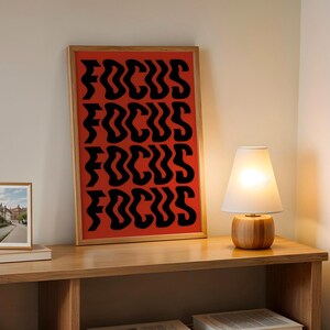 Könnte beinhalten: Ein gerahmter Kunstdruck mit dem Wort "FOCUS" in Schwarz auf rotem Hintergrund. Ein kleines gerahmtes Foto und eine Holzlampe mit weißem Schirm stehen auf einem Holzregal.