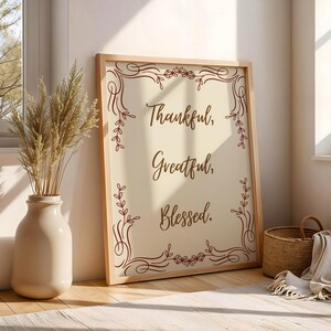 Könnte beinhalten: Ein rechteckiges gerahmtes Wandbild mit cremefarbenem Hintergrund und dunkelbraunem Text, der Thankful, Greatful, Blessed lautet. Der Druck zeigt einen dunkelbraunen dekorativen Rand mit geschwungenen Linien und kleinen Blattmotiven.