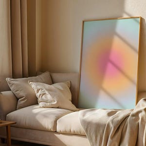 Puede incluir: Una impresi&oacute;n art&iacute;stica abstracta enmarcada con un degradado de colores, incluyendo rosa, naranja y azul, se apoya en un sof&aacute; beige con cojines y una manta. La habitaci&oacute;n tiene cortinas beige y una mesa auxiliar de madera.
