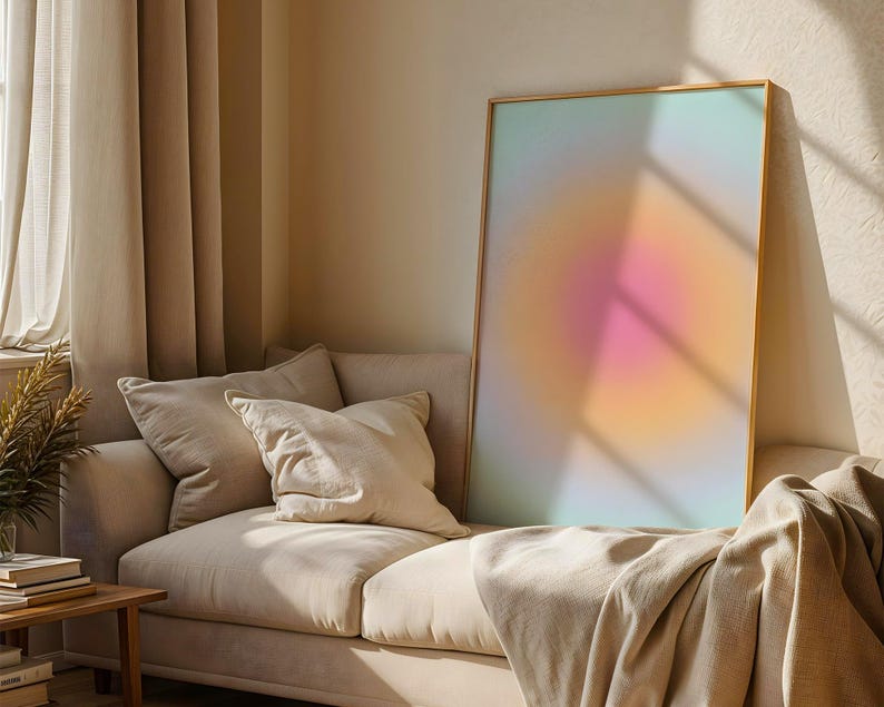 Puede incluir: Una impresi&oacute;n art&iacute;stica abstracta enmarcada con un degradado de colores, incluyendo rosa, naranja y azul, se apoya en un sof&aacute; beige con cojines y una manta. La habitaci&oacute;n tiene cortinas beige y una mesa auxiliar de madera.