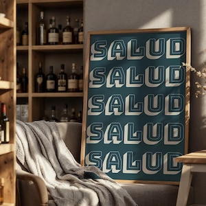 May include: A framed teal poster with the word "SALUD" repeated five times in a 3D white font with a dark outline. The poster is displayed in a room with a wooden shelf filled with bottles and a cozy chair.