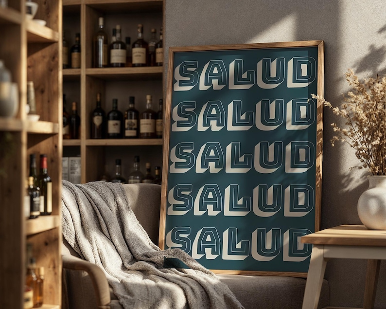 May include: A framed teal poster with the word "SALUD" repeated five times in a 3D white font with a dark outline. The poster is displayed in a room with a wooden shelf filled with bottles and a cozy chair.