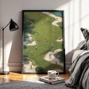 May include: A framed golf course art print. The artwork shows a green golf course with sand traps, water, and golfers. The print is displayed on a wooden floor, next to a bed with a grey blanket and pillow. A black lamp and a coffee cup are also visible.