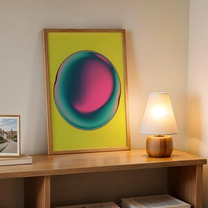 Könnte beinhalten: Ein gerahmter Kunstdruck mit einem leuchtenden rosa und türkis Design auf gelbem Hintergrund. Eine kleine gerahmte Fotografie und eine Holzlampe mit weißem Schirm stehen auf einem Holzregal.