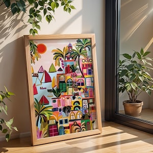 May include: A framed art print depicting a colorful village scene with palm trees, boats, and a setting sun. The artwork features a variety of architectural details and vibrant colors, including pink, blue, and green. The wooden frame is light-colored.