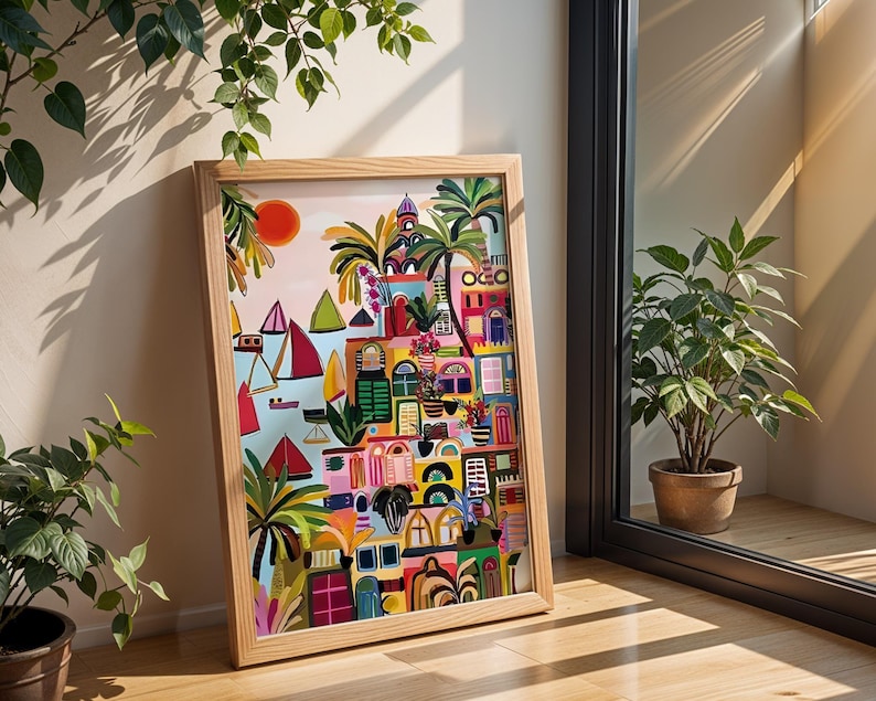 May include: A framed art print depicting a colorful village scene with palm trees, boats, and a setting sun. The artwork features a variety of architectural details and vibrant colors, including pink, blue, and green. The wooden frame is light-colored.