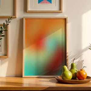 May include: A framed abstract art print with a gradient of orange, red, and teal hues. The artwork is displayed on a wooden surface, alongside a wooden plate holding three pears and a small orange fruit. Decorative plants in pots are also present.