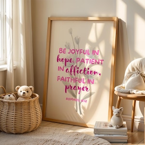 Könnte beinhalten: Gerahmter Druck mit den Worten „BE JOYFUL in hope, PATIENT in affliction, FAITHFUL in prayer“ in rosa Schrift. Ein Weidenkorb mit Teddybären, ein Stuhl mit einer Decke und ein kleines Stofftier befinden sich im Raum.