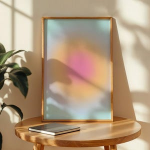 Puede incluir: Una impresi&oacute;n art&iacute;stica enmarcada con un degradado suave y colorido de tonos rosa, naranja y azul. La obra de arte se muestra en un marco de madera natural y descansa sobre una mesa redonda de madera. Un cuaderno cerrado est&aacute; sobre la mesa.