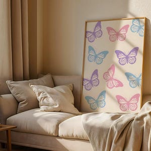 Könnte beinhalten: Ein gerahmter Druck mit einem Muster aus stilisierten Schmetterlingen in Lila, Rosa und Blau auf cremefarbenem Hintergrund. Das Kunstwerk wird auf einem beigen Sofa mit Kissen und einer Decke präsentiert. Der Raum hat beige Vorhänge und einen hölzernen Beistelltisch.