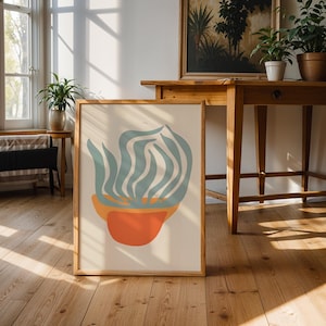 Könnte beinhalten: Ein gerahmter Kunstdruck mit einem abstrakten Pflanzendesign. Das Kunstwerk zeigt einen rot-orangen Topf und blau-grüne Pflanzenformen auf einem hellbeigen Hintergrund. Der Druck steht auf einem Holzboden und lehnt an einer Wand.