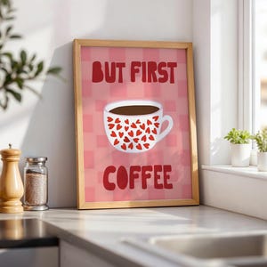 Könnte beinhalten: Ein gerahmter Kunstdruck mit einem rosa und hellrosa karierten Hintergrund und einer weißen Kaffeetasse, die mit kleinen roten Herzen verziert ist. Der Text BUT FIRST COFFEE ist in fetten roten Buchstaben über und unter der Tasse abgebildet.