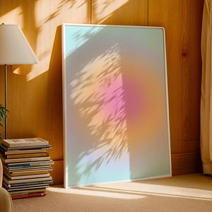 Puede incluir: Una impresi&oacute;n art&iacute;stica enmarcada con un degradado suave y colorido de tonos rosa, naranja y azul. La obra de arte est&aacute; apoyada contra una pared de madera, con sombras de hojas proyectadas sobre ella. Una l&aacute;mpara y una pila de libros est&aacute;n en primer plano.