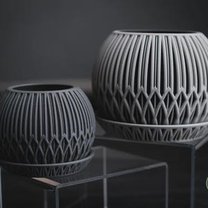 Op de afbeelding: Twee grijze, bolvormige plantenbakken met ingewikkelde verticale en ruitvormige patronen. Elke plantenbak staat op een bijpassende schotel. De plantenbakken worden tentoongesteld op een heldere, moderne standaard tegen een donkere achtergrond.