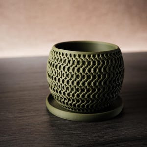 Könnte beinhalten: Olivgrüner Keramik-Pflanztopf mit passender Untertasse. Der Topf hat ein einzigartiges, offenes Design mit einem sich wiederholenden Wellenmuster. Der runde Topf steht auf einem kleinen, kreisförmigen Sockel. Der Hintergrund ist ein neutraler, verschwommener Farbverlauf.