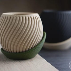 Könnte beinhalten: Zwei dekorative Pflanzgefäße mit Spiralmuster. Eines ist hellbeige, das andere schwarz. Beide stehen auf einer grünen Untertasse. Die Pflanzgefäße sind wahrscheinlich für kleine Pflanzen oder Sukkulenten.