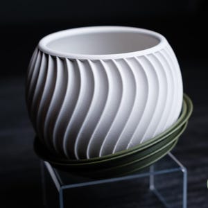Pode incluir: Um vaso branco, esférico, com um design texturizado em forma de onda. O vaso está sobre um pires verde escuro. O design é de h3li0 design.