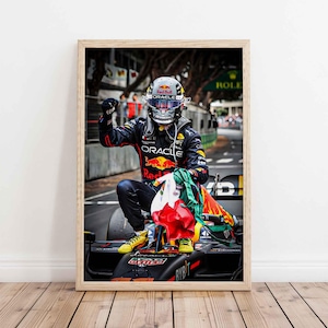 May include: Framed print of a Formula 1 driver celebrating a victory. The driver is wearing a helmet and racing suit with sponsor logos, kneeling on the car. The image includes a flag and the Red Bull and Oracle logos.