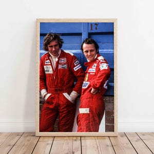 May include: Framed print featuring two men in red racing suits, standing in front of a blue wall. The men are smiling, and the print has a wooden frame. The suits have sponsor logos.