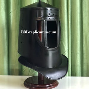 May include: A black metal helmet with a visor and a decorative black feather plume. The helmet is displayed on a wooden stand. The text "RM-replicamuseum" is visible on the helmet.