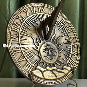 May include: A bronze-colored sundial with Roman numerals and a sun face design. The sundial has a gnomon and is mounted on a decorative stand. The text "RM-replicamuseum" is visible on the bottom left of the sundial.