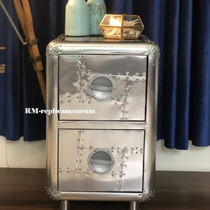 Può includere: Un mobile in metallo argentato con due cassetti, ciascuno con un design circolare a oblò. L'armadio presenta dettagli rivettati e poggia su gambe corte e cilindriche. Due bottiglie azzurre e un portacandele geometrico in vetro sono sopra.