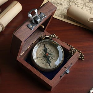 Puede incluir: Una brújula vintage en una caja de madera con una cadena de latón. La brújula tiene una esfera detallada con marcas direccionales. La caja está abierta, revelando la brújula contra un forro de terciopelo azul. Pergaminos enrollados y un mapa en el fondo.