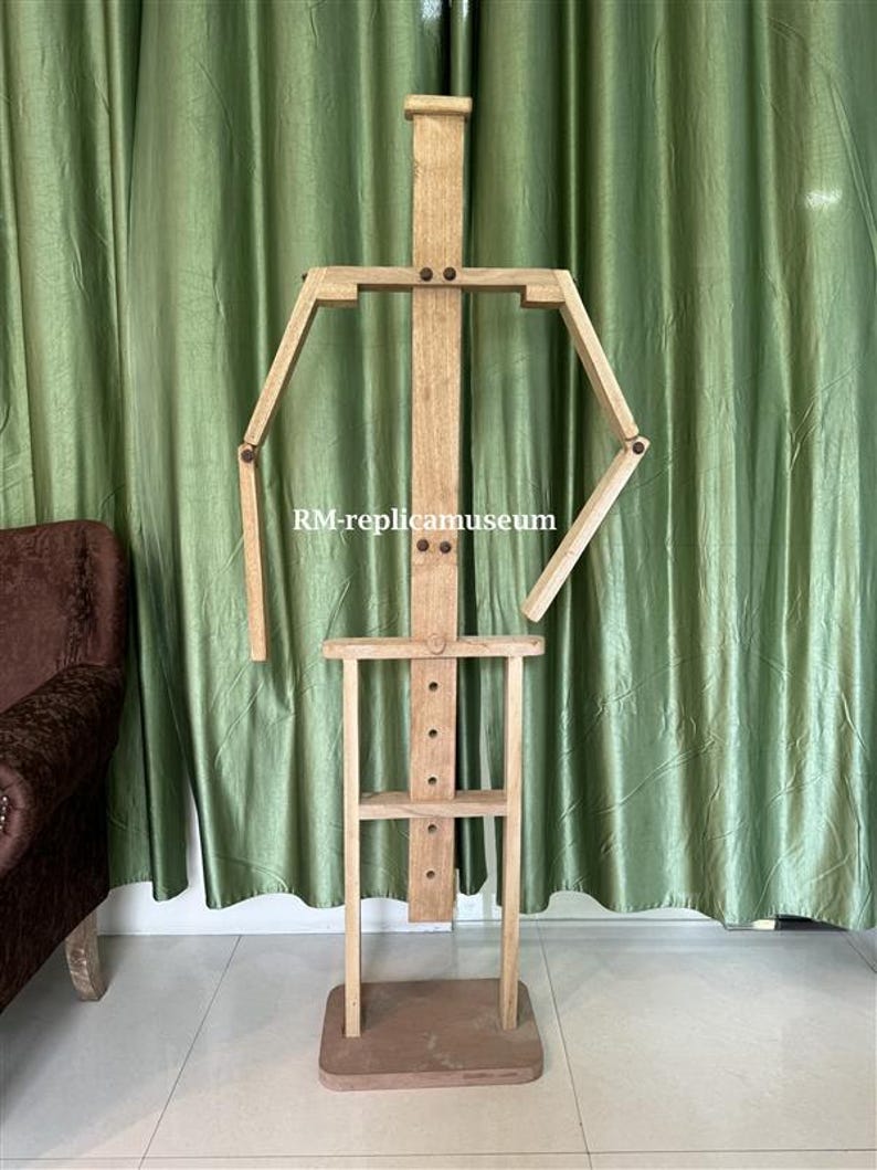 May include: A wooden artist's mannequin with articulated arms and legs, standing on a rectangular base. The mannequin is made of light-colored wood and features a simple, geometric design. The text "RM-replicamuseum" is visible on the mannequin.