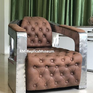 May include: A brown leather armchair with a button-tufted design. The chair has a metal frame with riveted details and padded armrests. The text "RM-ReplicaMuseum" is visible on the front of the chair.