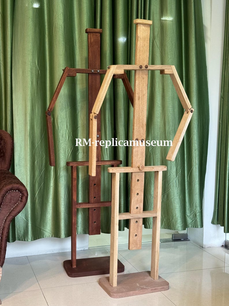 May include: Two wooden artist mannequins. One is a light brown color, and the other is a darker brown. The mannequins have articulated arms and legs, and are standing in front of a green curtain. The text "RM-replicamuseum" is visible.