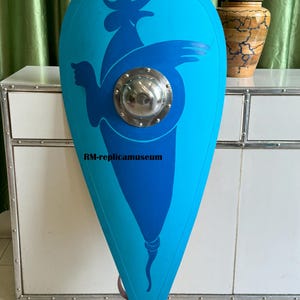 May include: A light blue, teardrop-shaped shield with a dark blue bird design and a silver metal boss. The shield has a rounded top and a pointed bottom. The text "RM-replicamuseum" is printed on the shield.