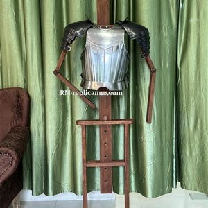 May include: A medieval-style metal breastplate and shoulder armor displayed on a wooden mannequin. The armor is silver with black accents, and the mannequin has wooden arms and a dark brown base. The text "RM-replicamuseum" is visible.