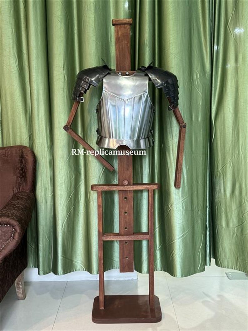 May include: A medieval-style metal breastplate and shoulder armor displayed on a wooden mannequin. The armor is silver with black accents, and the mannequin has wooden arms and a dark brown base. The text "RM-replicamuseum" is visible.