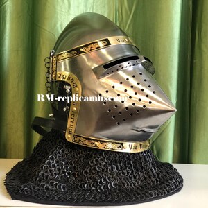 May include: A silver-colored medieval helmet with a pointed visor and numerous small ventilation holes. The helmet features gold-colored decorative bands with ornate designs and text. A chainmail neck guard is attached. The helmet is displayed on a white surface.