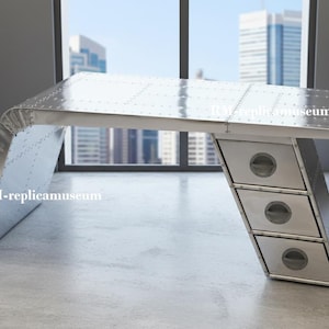 May include: A silver metal desk with a riveted design, featuring a slanted leg and three drawers with circular handles. The desk has a modern, industrial aesthetic. The desk is in front of a window with a city view.