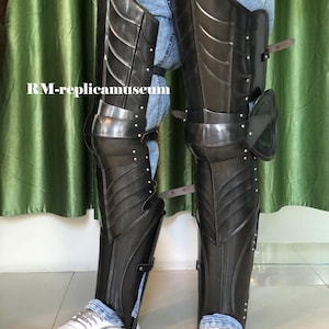 May include: Black metal leg armor with riveted accents and articulated plates. The armor is worn over blue jeans and white sneakers. The text "RM-replicamuseum" is visible on the left side of the image.