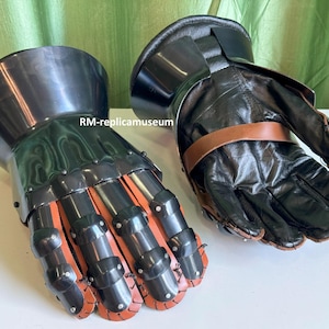 May include: Pair of black and silver medieval gauntlets. The gauntlets feature articulated metal plates over the knuckles and fingers, with red leather accents. The palms and cuffs are made of black leather, secured with brown straps.