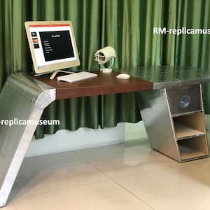 May include: A silver metal desk with a brown wooden top and riveted metal accents. The desk has a built-in drawer and two shelves. A computer monitor, keyboard, and mouse sit on the desk. The desk is in front of a green curtain.