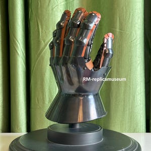 May include: A black and brown metal gauntlet, a type of medieval armor, is displayed on a black stand. The gauntlet features articulated fingers and a flared wrist. The text "RM-replicamuseum" is visible on the image.