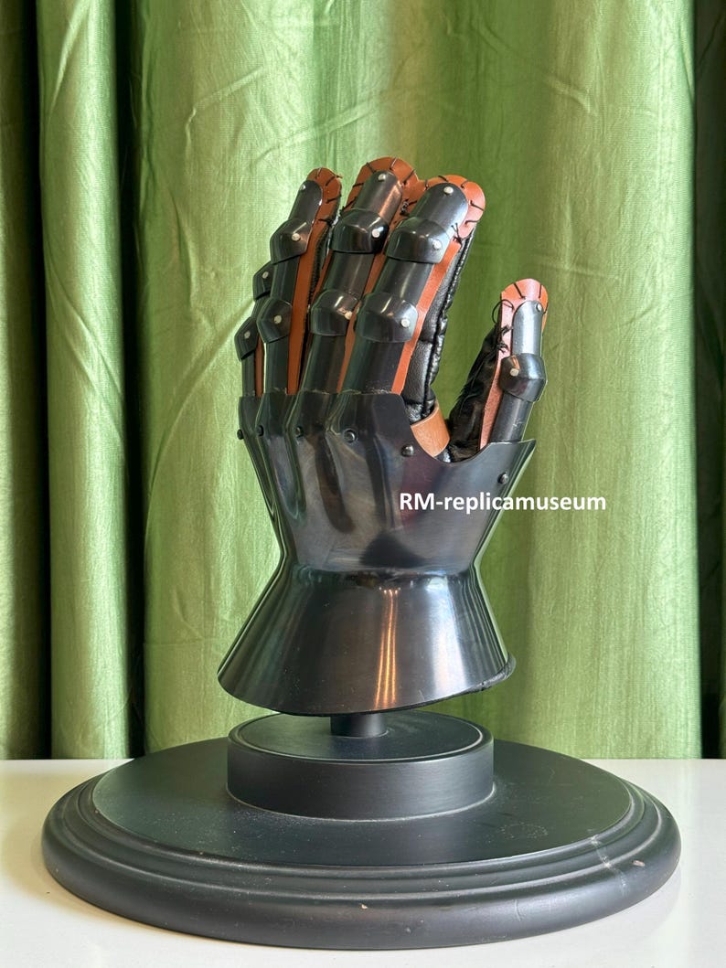 May include: A black and brown metal gauntlet, a type of medieval armor, is displayed on a black stand. The gauntlet features articulated fingers and a flared wrist. The text "RM-replicamuseum" is visible on the image.
