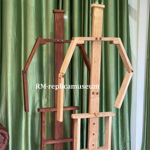 May include: Two wooden artist easels. One is a dark brown color, and the other is a light natural wood color. Both easels have adjustable arms and a base for holding a canvas. The text "RM-replicamuseum" is visible.