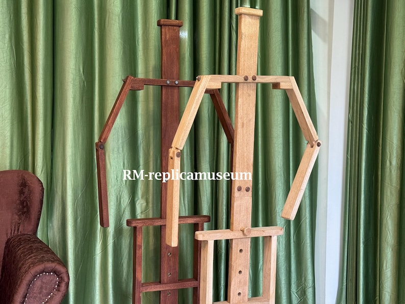 May include: Two wooden artist easels. One is a dark brown color, and the other is a light natural wood color. Both easels have adjustable arms and a base for holding a canvas. The text "RM-replicamuseum" is visible.
